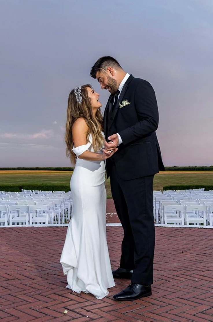 Bride in an off-shoulder fit and flare wedding dress, faces her groom and they dance together at an outdoor ceremony.