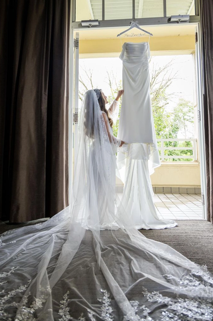 Bride looks up at an off-shoulder draped satin bodice gown, hanging from the top of a doorway with a long lace-edge veil.