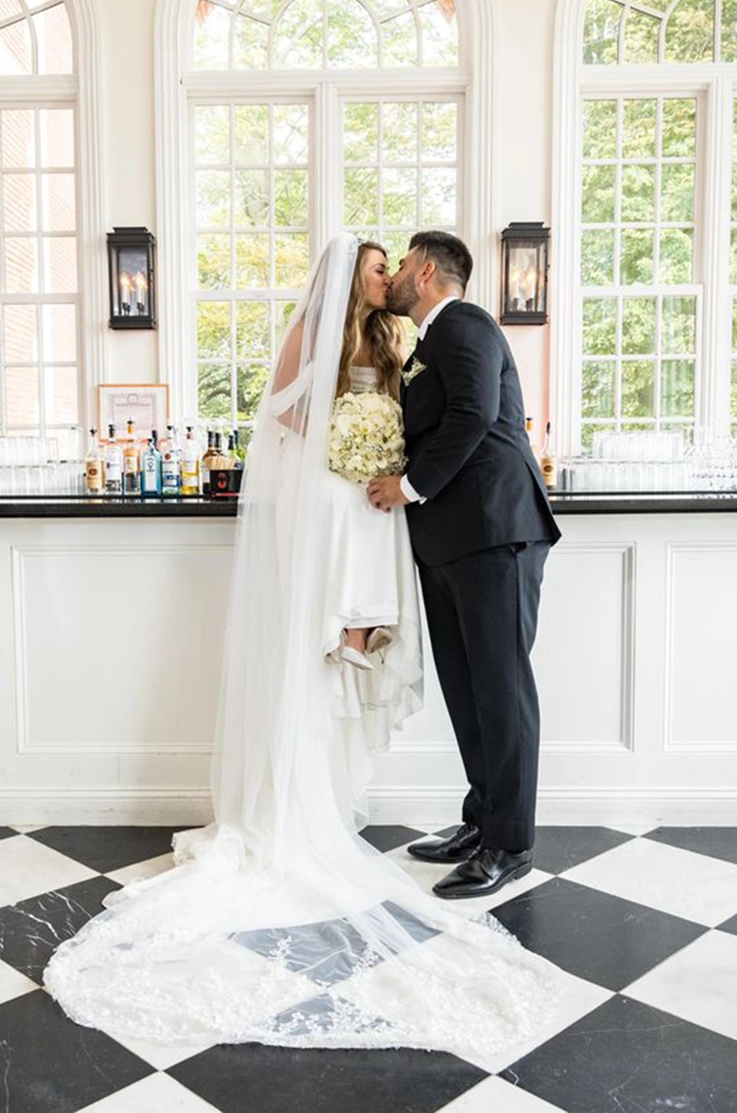 Groom kisses bride sitting on a bar countertop in a charmeuse satin gown with draped sleeves and a veil, holding a bouquet.