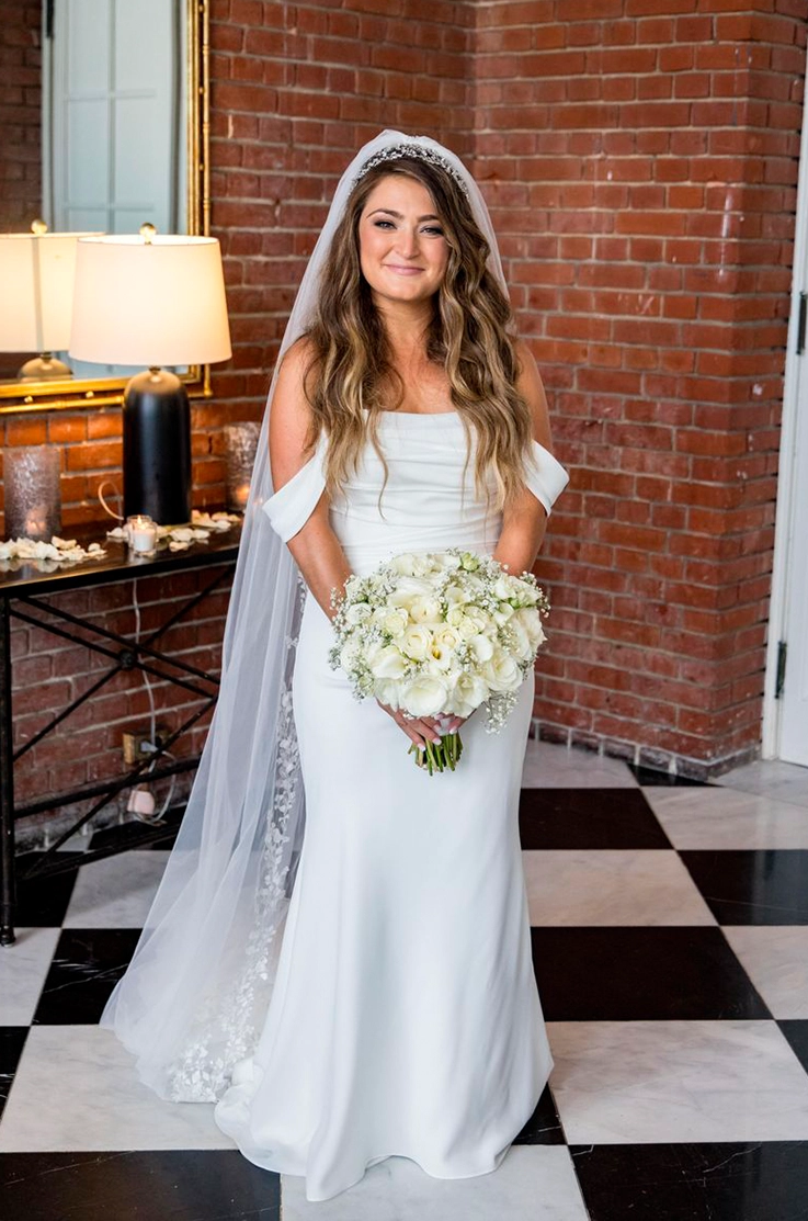 Smiling bride in a satin fit and flare gown with draped sleeves and a lace-trim veil, holding a white rose bouquet indoors.