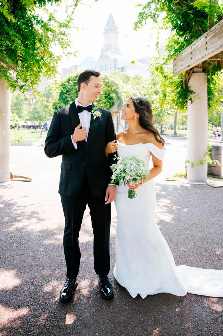 Bride in a fit and flare satin wedding gown with draped sleeves, faces her groom in an urban city park between trees.