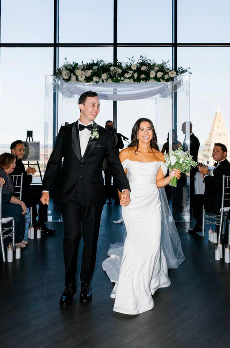 Bride in a fitted charmeuse satin wedding dress with a draped bodice, walks down the aisle with her groom in a modern venue.