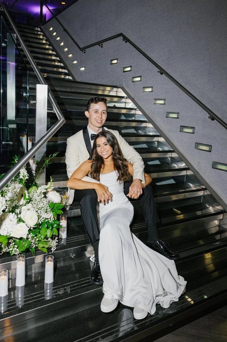 Bride in a strapless fit and flare gown with draping, poses with groom on a modern black staircase with candles.