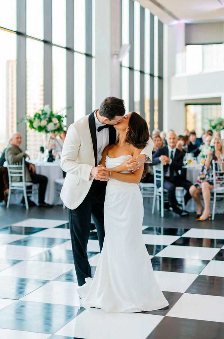 Bride in a scoop neck satin gown with draping, kisses groom in an embrace on a checkered dance floor in a glass building.