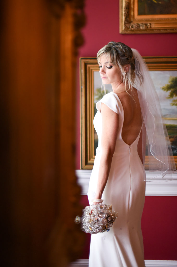 A bride in a cap sleeve wedding dress and veil holds a bouquet and looks over her shoulder, while standing indoors with art on the wall behind her.
