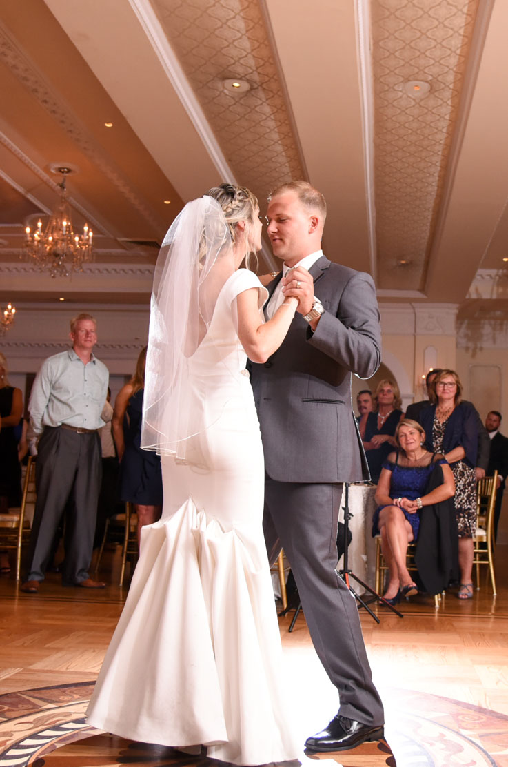 A bride in a bustled cap sleeve wedding dress and veil, and a groom in a suit, dance together in a ball room as wedding guests watch in the background.