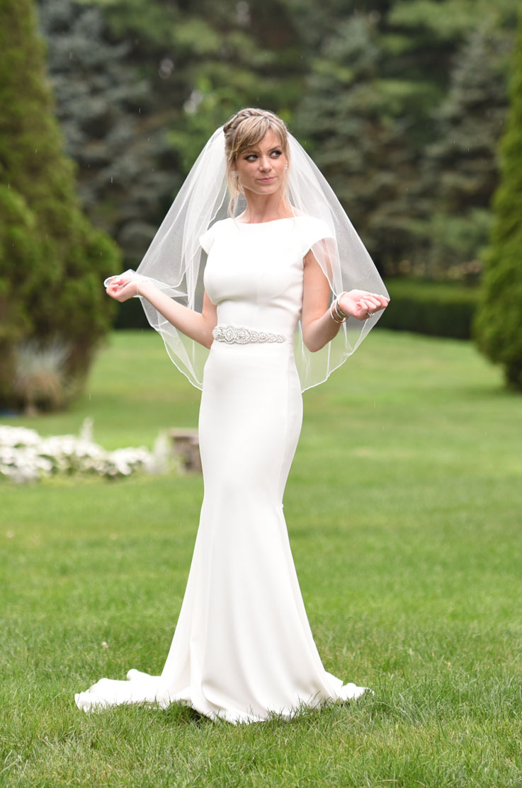 A bride in a cap sleeve wedding dress and veil stands outside in a grass field and looks off to the side, while she holds the edges of her veil up a bit.