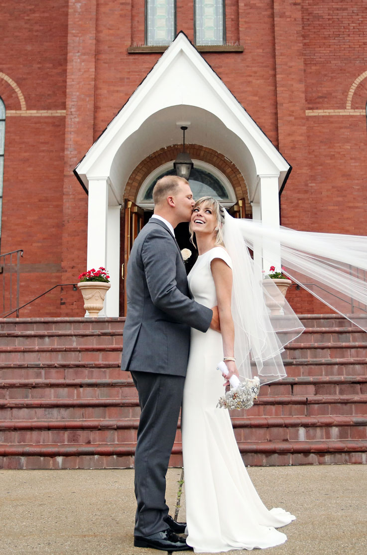 A groom in a suit kisses the head of a smiling bride in a cap sleeve wedding dress and veil, as they stand outside at the bottom of a brick staircase, in front of a building.