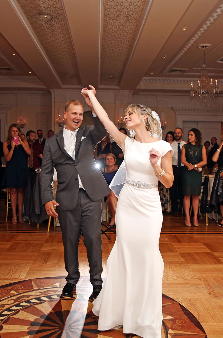 A groom in a suit spins a bride in a cap sleeve wedding dress and veil in a ball room, both of them smiling, as wedding guests watch in the background.