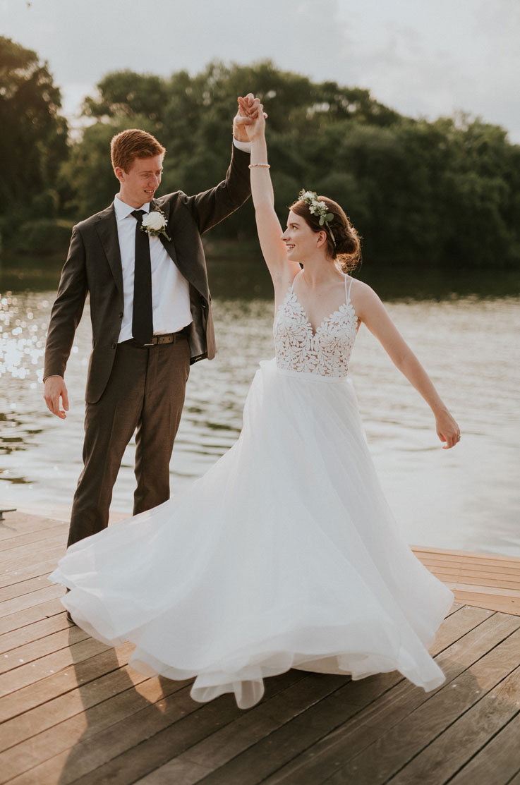 A groom in a suit spins a bride in an Organza ball gown wedding dress on a dock, with a lake and trees in the background.