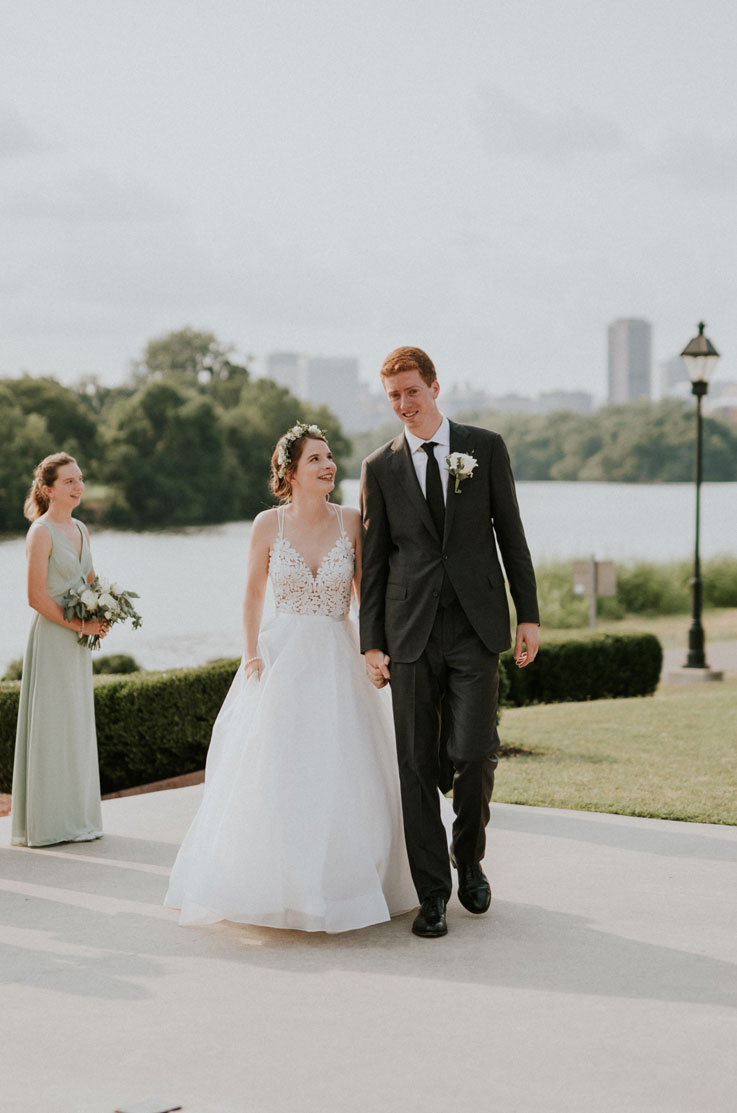 A bride in an Organza ball gown wedding dress and a groom in a suit walk hand in hand in a park, the bride is looking at the groom, with a bridesmaid, a lake, and buildings in the background.