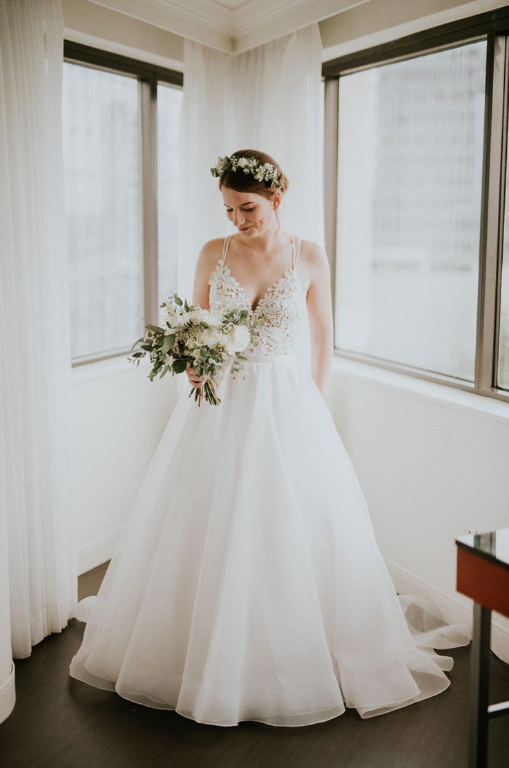 A bride in an Organza ball gown wedding dress stands indoors while holding a bouquet, with windows and curtains behind her.