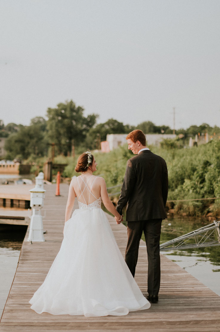 A bride in an Organza ball gown wedding dress and a groom in a suit look at each other and walk hand in hand down a dock on a lake, with trees and buildings in the background.