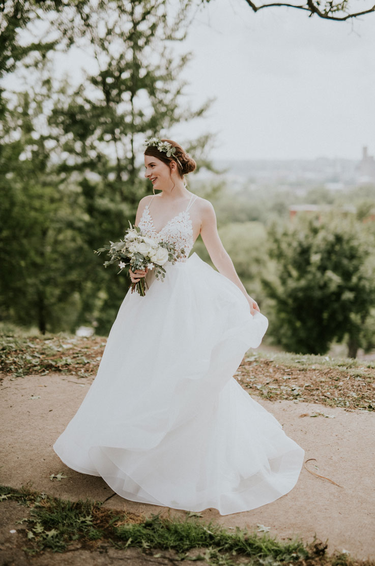 A bride in an Organza ball gown wedding dress stands on a path outdoors in a park, holding a bouquet and looking off to the side.