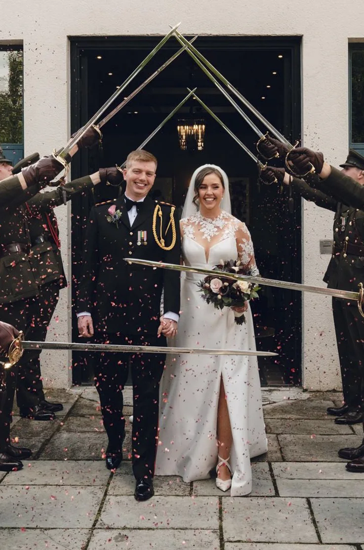 Bride in a Mikado illusion neck gown with sleeves, skirt slit and a veil, walks beside groom in uniform through a sword arch.