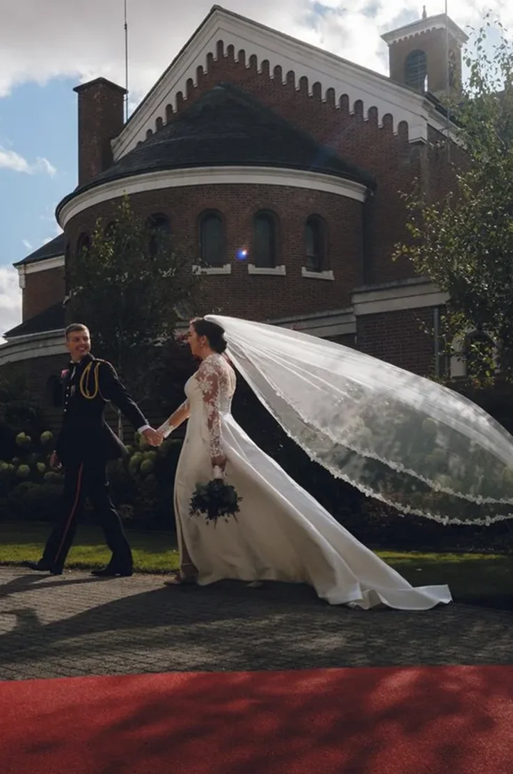 Bride in a lace sleeved Mikado wedding dress with a flowing veil, walks hand in hand with groom outside the venue.