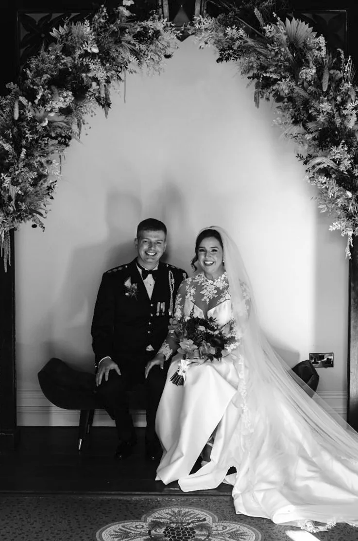 Bride in an illusion high neck gown with sheer lace sleeves and a veil, is sitting under a floral arch with groom, in black and white. 