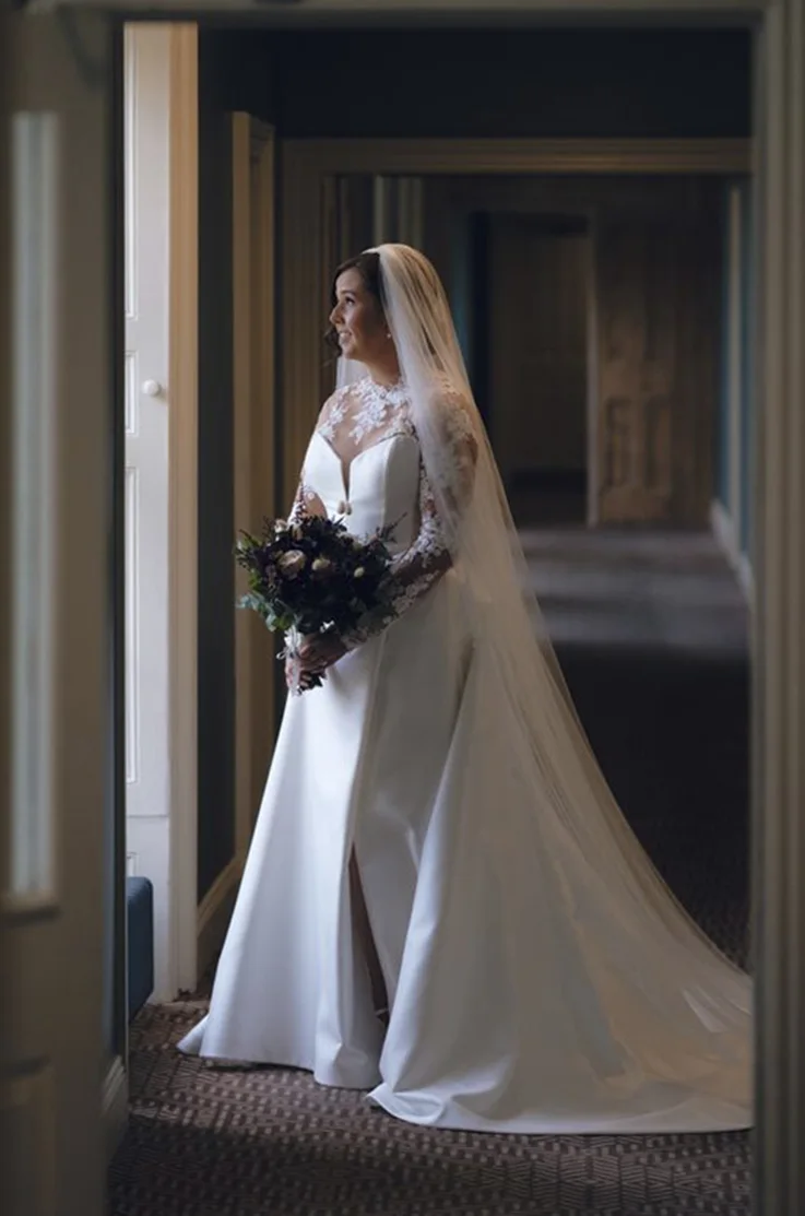 Bride poses in a doorway in an illusion neck and lace sleeved gown in Mikado with a veil, and is holding a bouquet.
