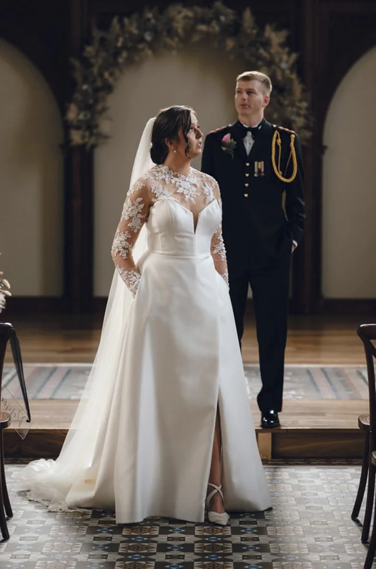 Bride in a Mikado A-line gown with a skirt slit, veil and an illusion lace neckline with sleeves, poses beside groom.