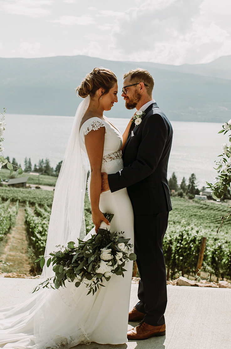 A bride in a fitted wedding dress and veil, holding a bouquet, stands facing a groom in a suit while he holds her, both of them standing outside in front of a vineyard with a lake and mountains in the background.