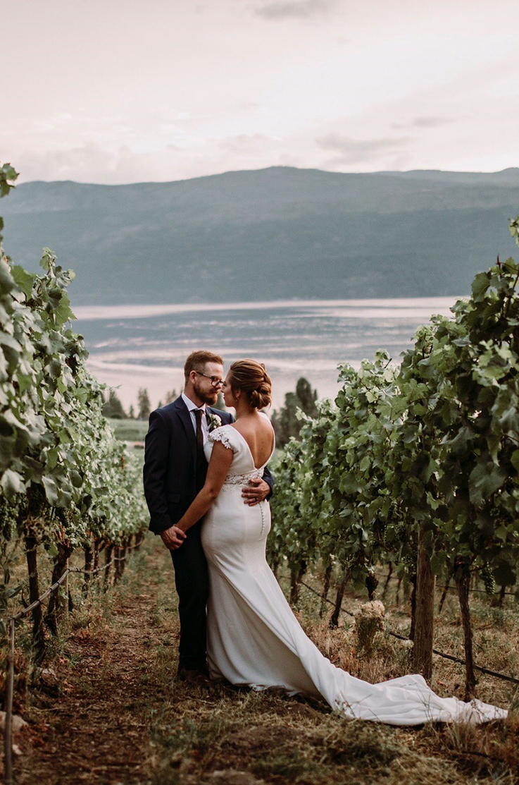 A bride in a fitted wedding dress and a groom in a suit hold each other while standing in a vineyard, with a lake and mountains in the background.