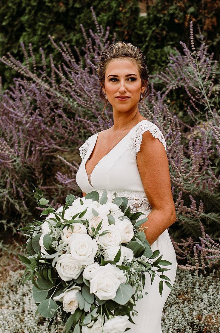 A bride in a cap sleeve wedding dress holds a bouquet and smiles while looking off to the side and standing outside in front of a garden.
