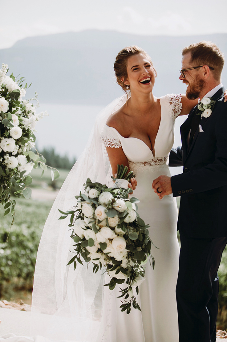 A bride in a fitted wedding dress and veil holds a bouquet, while laughing with a groom in a suit, both standing outside with mountains in the background.
