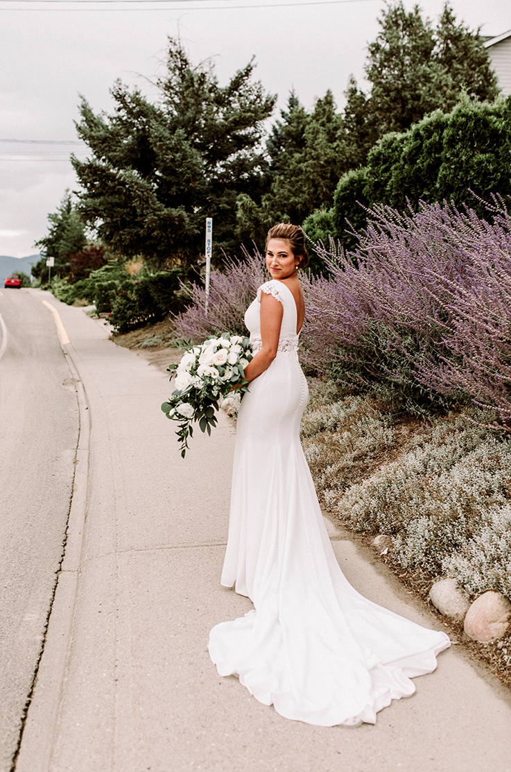 A bride in a fitted wedding dress holds a bouquet while smiling and looking over her shoulder, standing on a sidewalk in front of flowering bushes and trees.