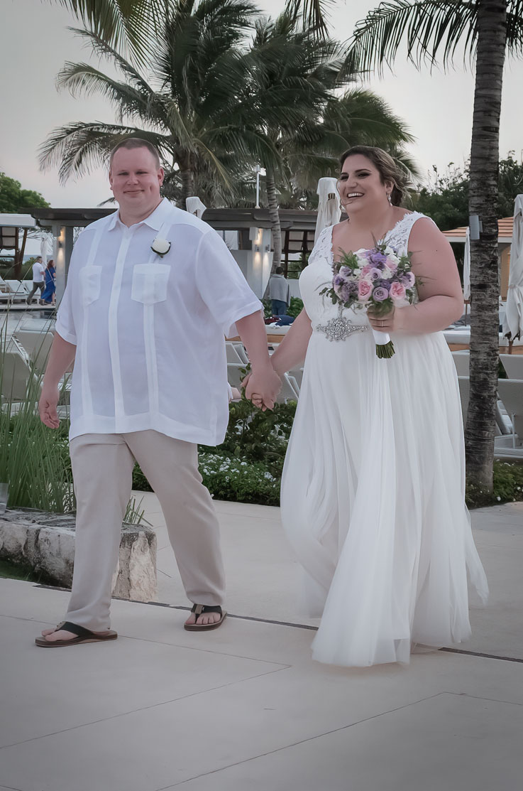 A smiling bride in a tulle skirt wedding dress holds a veil and walks with a groom in a short sleeve dress shirt at a resort, surrounded by palm trees.