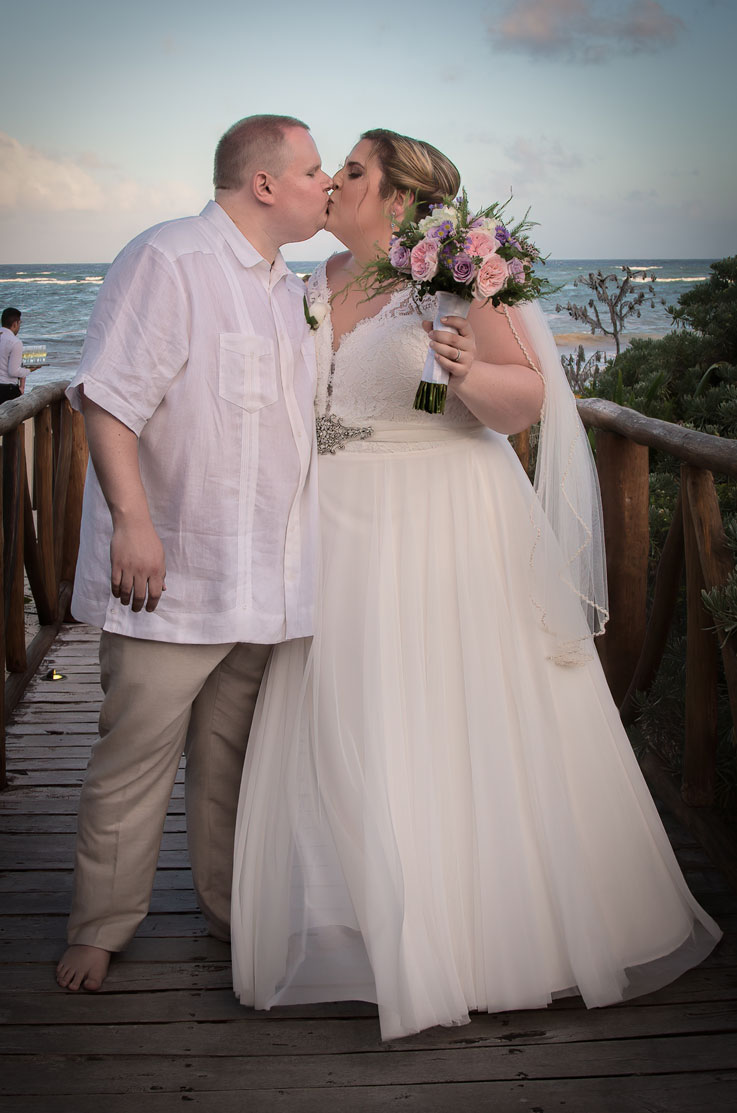 A bride in a tulle skirt wedding dress and a veil, holds a bouquet while kissing a groom in a short sleeve dress shirt as they stand on a pedestrian bridge in front of an ocean.