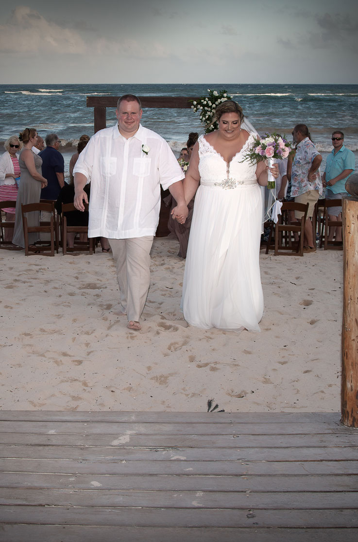 A bride in a tulle skirt wedding dress holding a veil and a groom in a short sleeve dress shirt walk up a beach with the ocean and wedding guests behind them.