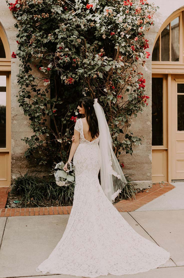 A bride in a cap sleeve lace wedding dress and veil holds a bouquet while standing outside in front of a flowering tree, with her lace train fanned out behind her.