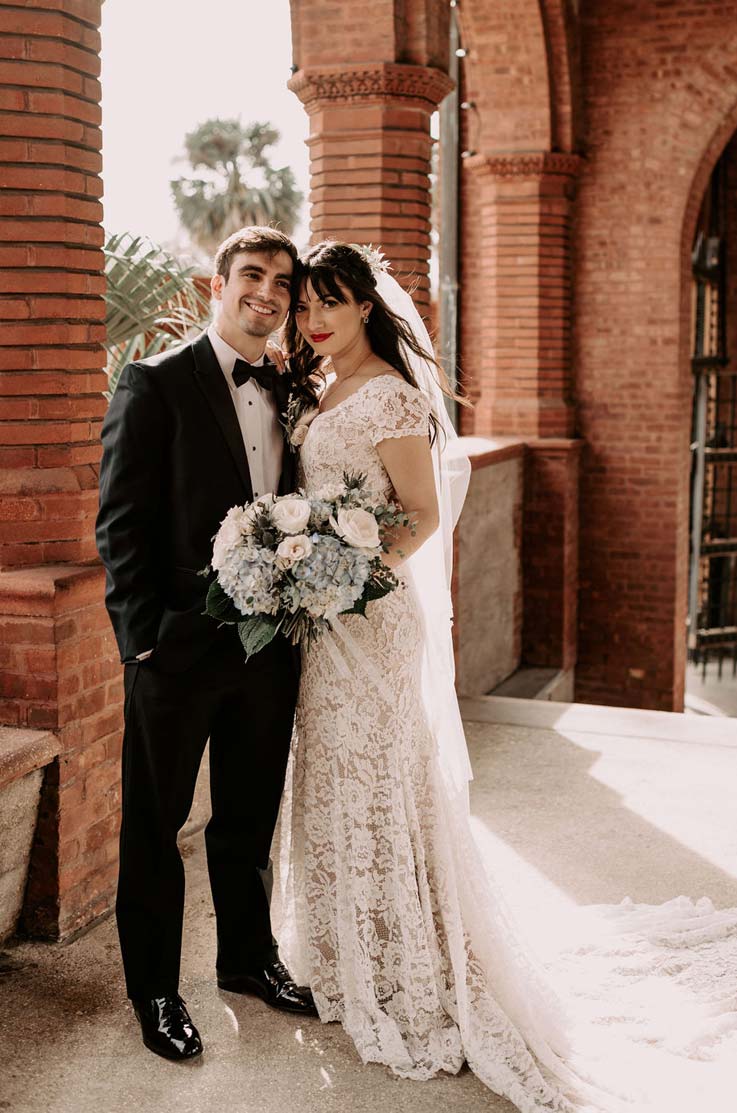 A bride in a cap sleeve lace wedding dress and veil, holding a bouquet, and a groom in a suit, stand beside each other with brick arches behind them.