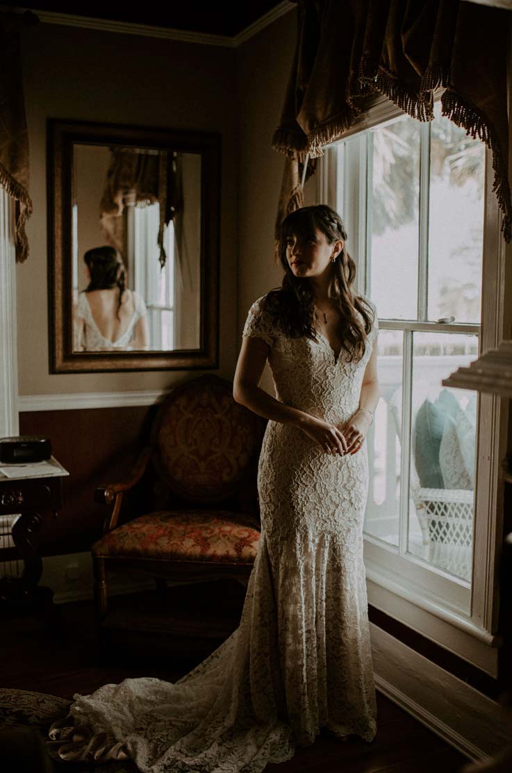 A bride in a cap sleeve lace wedding dress stands indoors in front of an ornate chair, mirror, and window while looking off to the side and smiling.