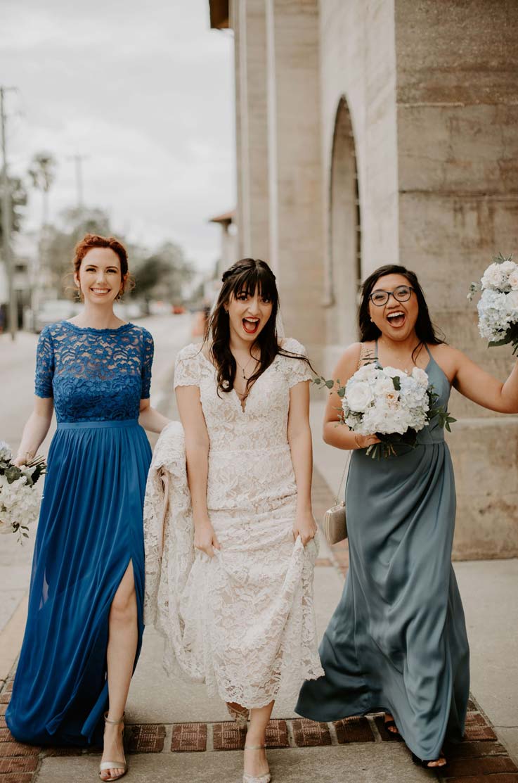 A bride in a cap sleeve lace wedding dress walks down the sidewalk with two bridesmaids, all of them smiling excitedly.