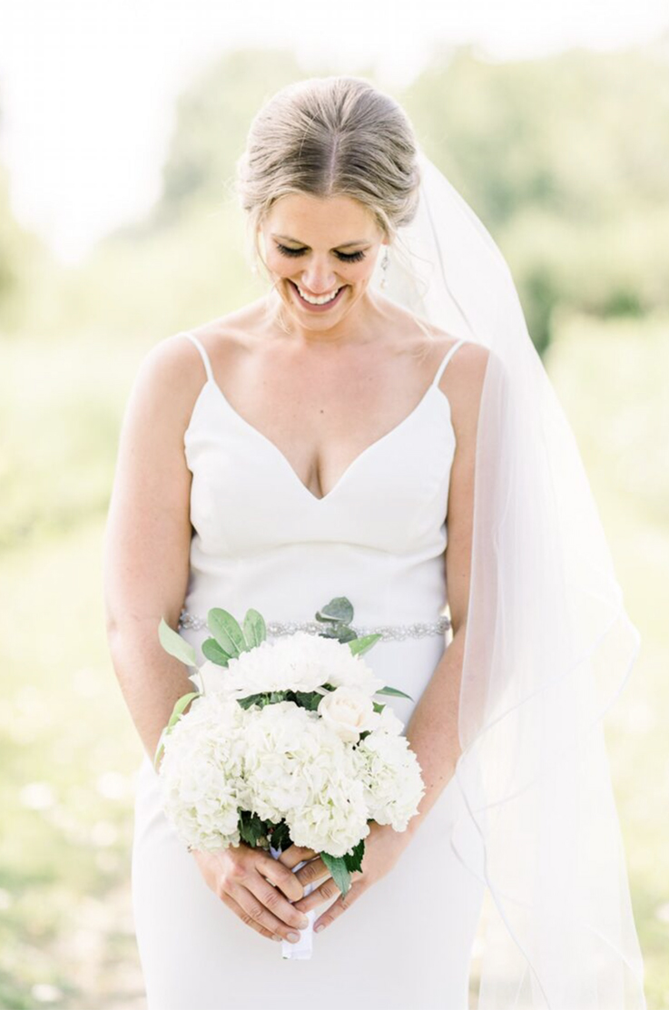 A bride in a V-neck wedding dress and veil holds a bouquet while smiling and looking down, while standing outside.