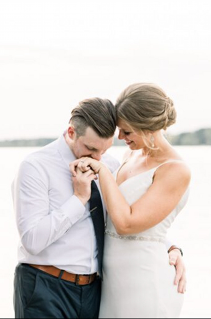 A groom in a dress shirt and tie kisses the hand of a bride in a V-neck wedding dress while she smiles, with a lake in the background.