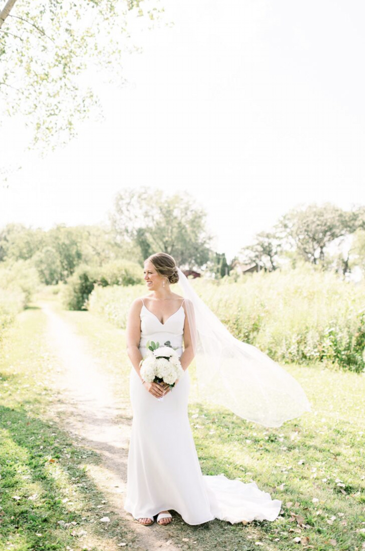 A bride in a V-neck wedding dress and veil holds a bouquet while smiling and looking off to the side, she is standing outside on path in a field.
