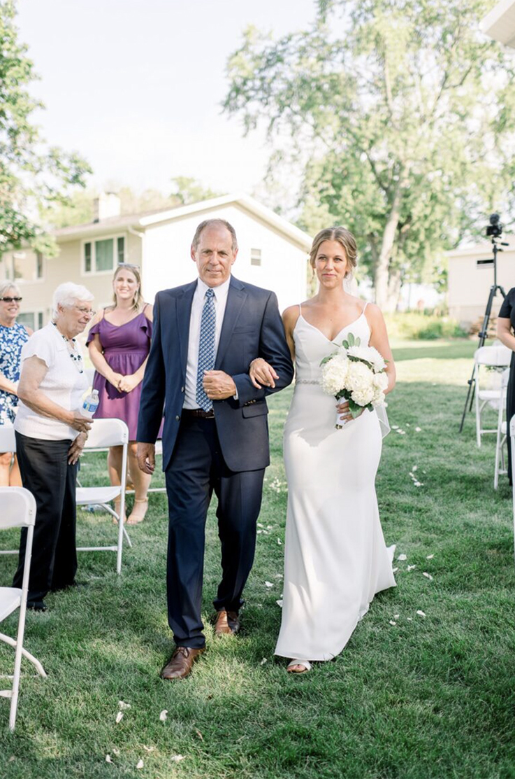 A bride in a V-neck wedding dress holds a bouquet while walking down an outdoor aisle with a man in a suit, with wedding guests standing to the side.