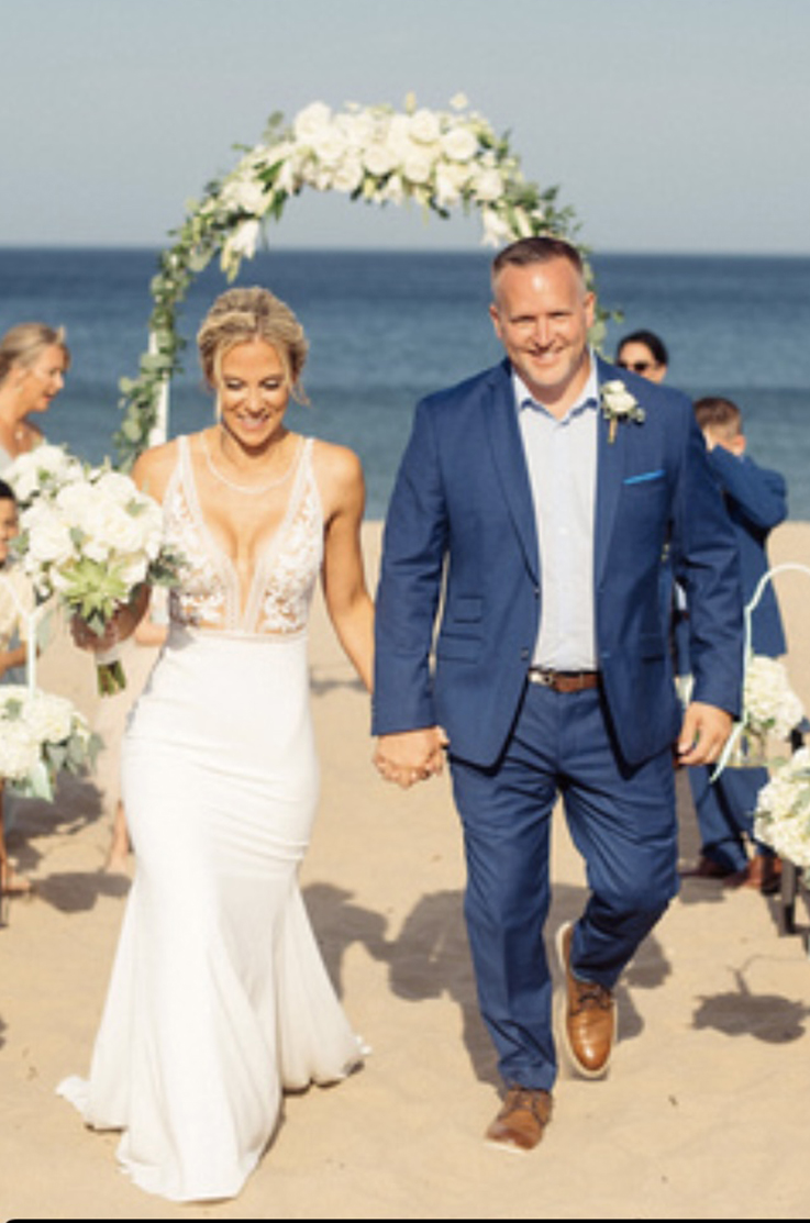 A bride in a plunging neckline wedding dress and a groom in a suit walk up a beach hand in hand while smiling, with a ceremony arch, wedding guests, and the ocean behind them. 