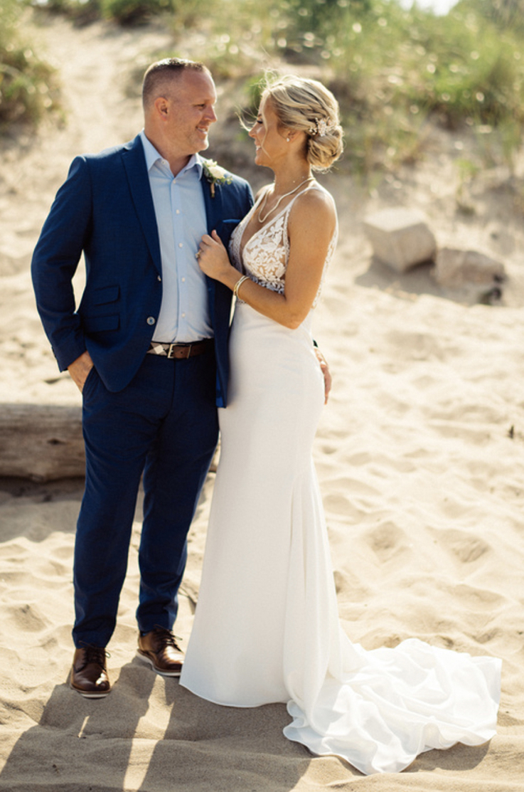 A bride in a plunging neckline wedding dress and a groom in a suit stand close and look at each other, smiling, while outside on a sandy beach.