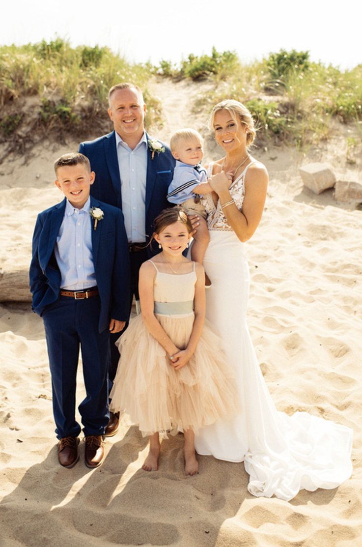 A bride in a plunging neckline wedding dress and a groom in a suit stand on a sandy beach with their three children.