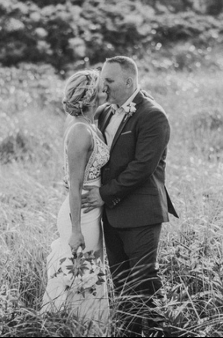 A bride in a sleeveless wedding dress holds a bouquet and kisses a groom in a suit, while they stand outside in a grassy field.