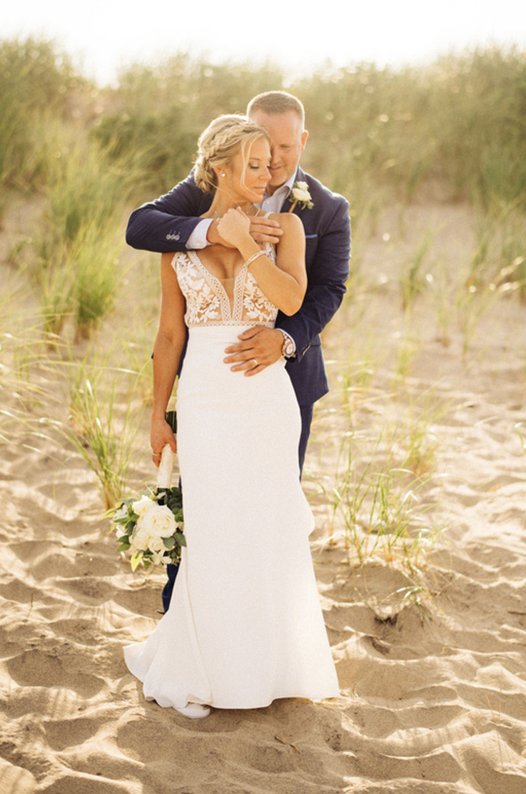 A groom in a suit stands behind a bride in a plunging neckline wedding dress and holds her, while they stand outside on a sandy beach.