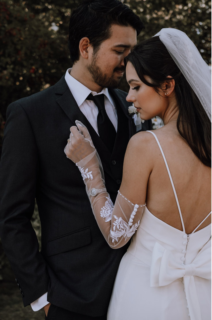 A bride in a fitted wedding dress, a veil, and lace sleeves stands close to a groom in a suit while they stand outdoors with trees in the background.