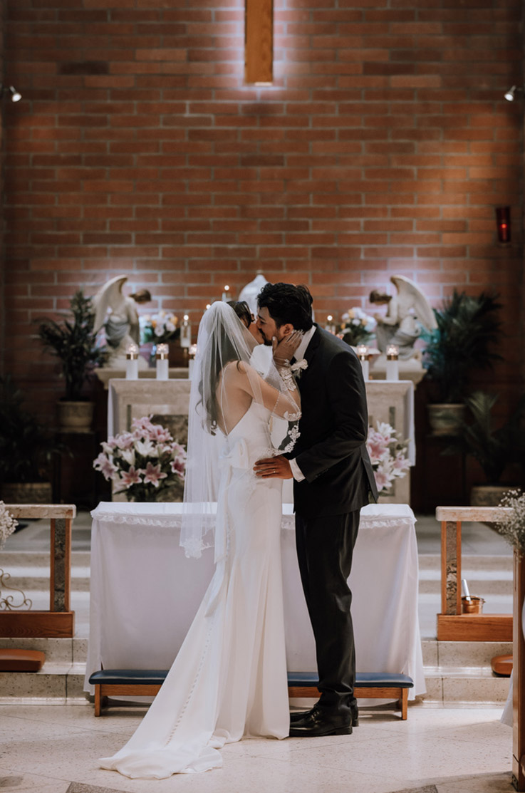 A bride in a fitted wedding dress and veil, and a groom in a suit kiss at the altar of a church.