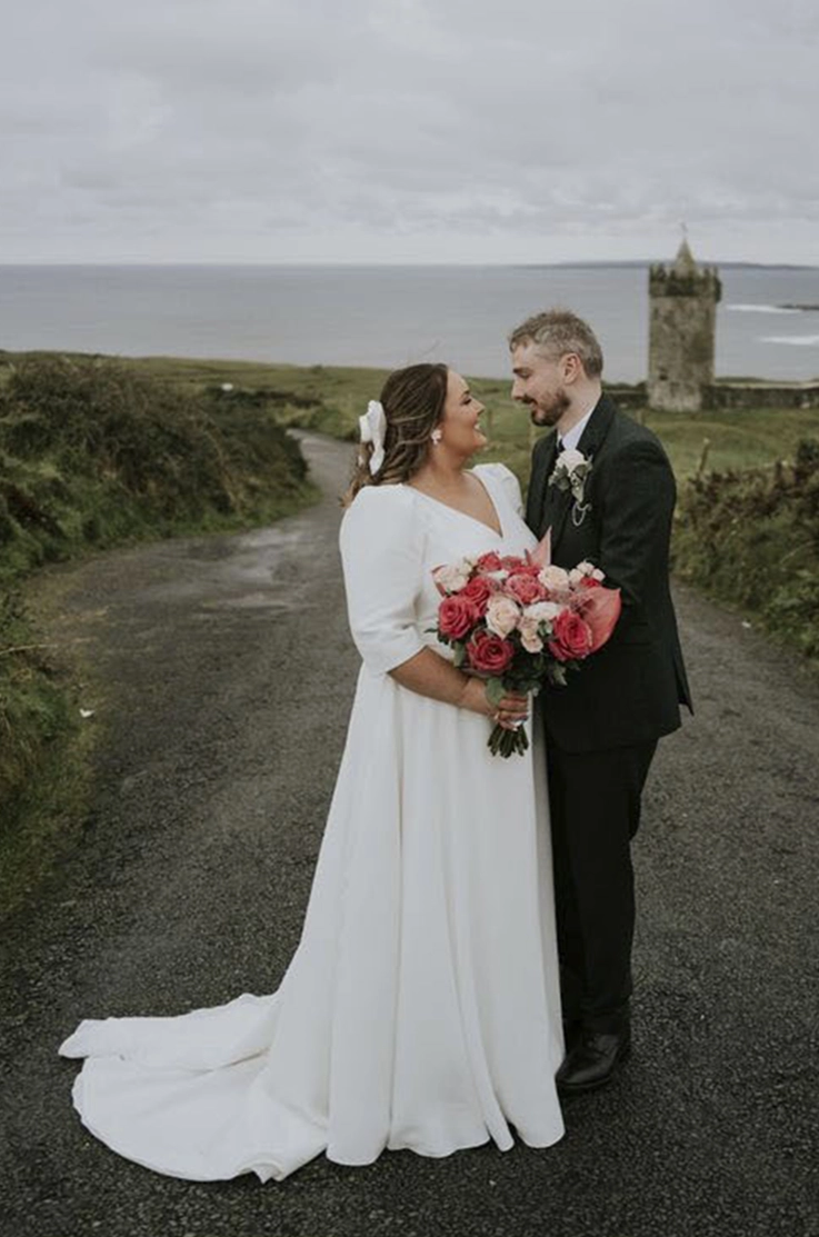Bride in a V-neck crepe gown with cap sleeves, poses with groom and holds a rose bouquet on an outdoor path near a castle.
