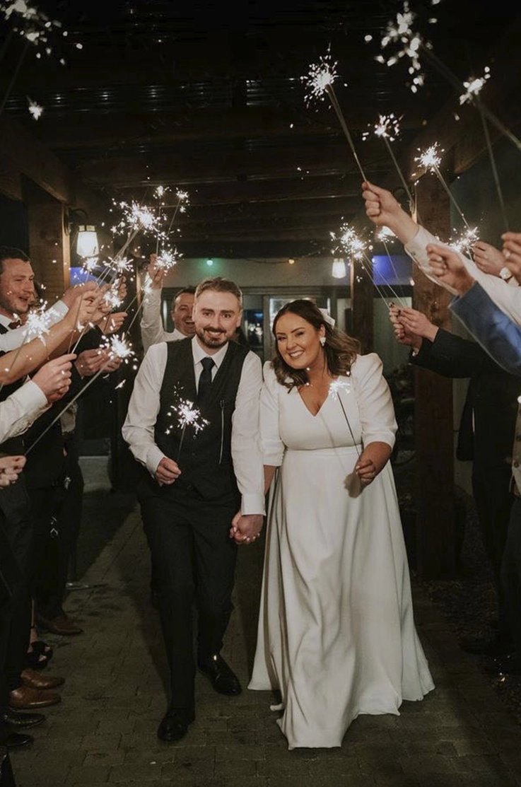 Bride in a crepe three-quarter sleeves gown with a V-neck, exits with her groom past sparklers. 
