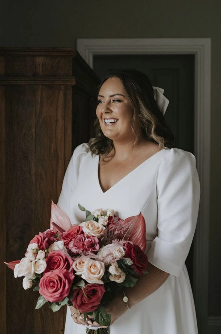 Close up of bride in a V-neck crepe wedding dress with mid length cap sleeves, and is holding a pink rose bouquet. 