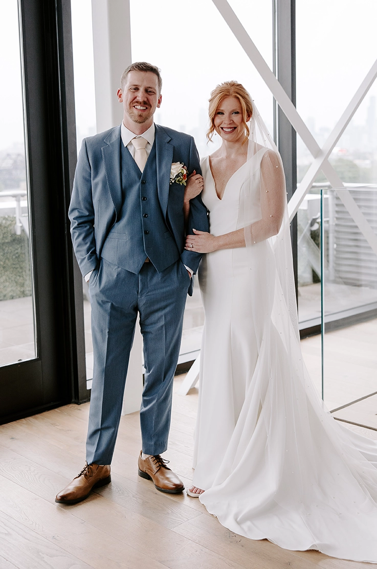Bride in a fit and flare crepe gown with a V-neckline and pearl veil, poses beside groom in a rooftop building.